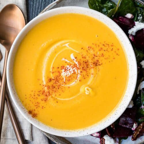 A bowl of creamy butternut squash soup garnished with a swirl of cream and sprinkled with paprika, served next to a salad on a plate with rose gold utensils.