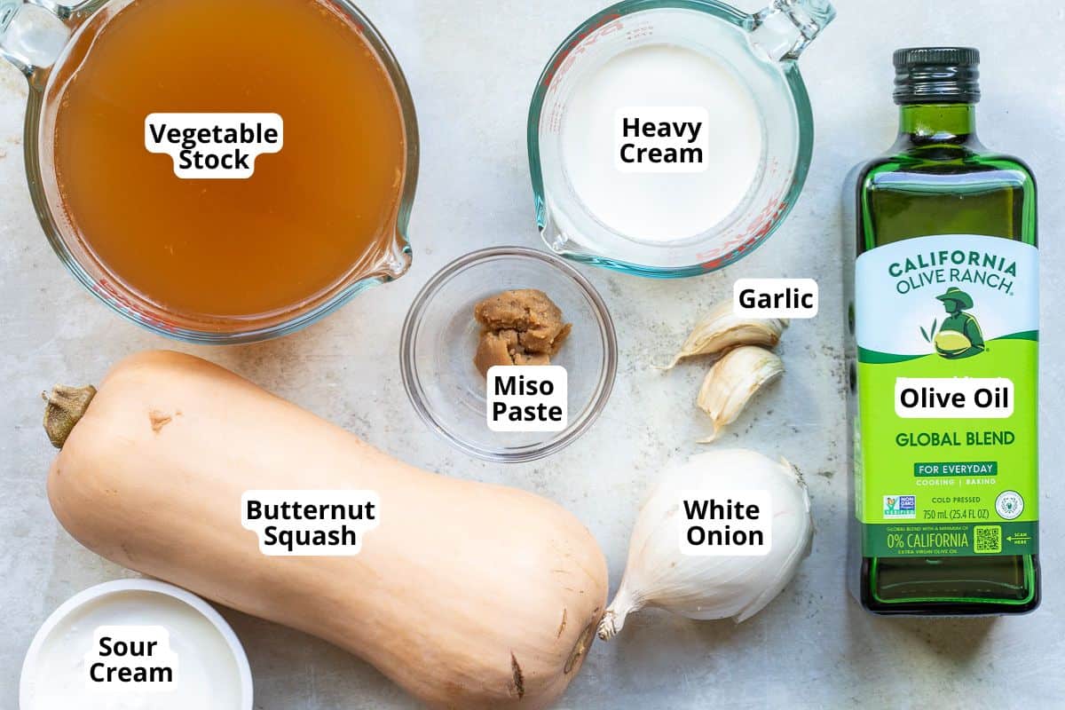 Ingredients for soup displayed on a counter: vegetable stock, heavy cream, olive oil, butternut squash, sour cream, white onion, garlic cloves, and miso paste.