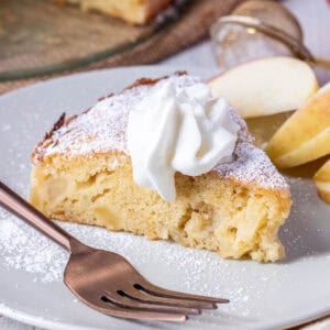A slice of apple cake topped with whipped cream and powdered sugar, served on a plate with apple slices and a fork.