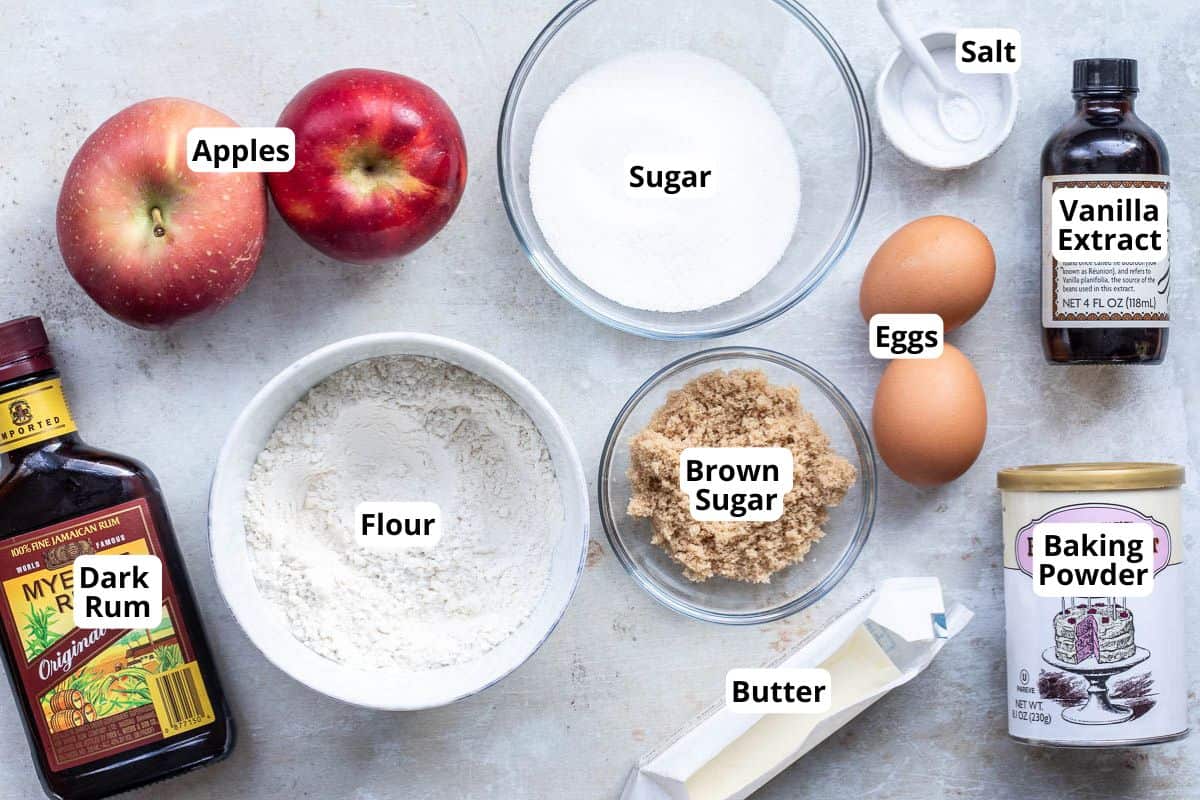 Top-down view of baking ingredients including apples, flour, sugar, eggs, butter, brown sugar, dark rum, baking powder, vanilla extract, and salt, each labeled.