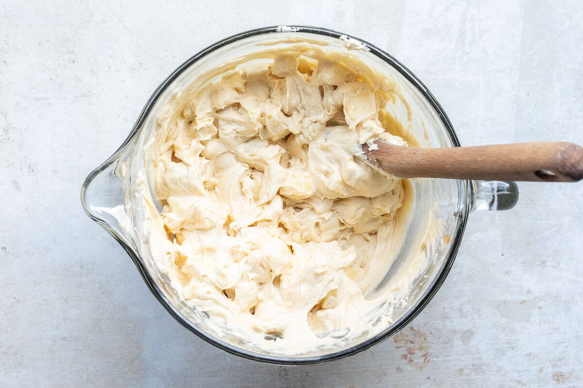 A glass mixing bowl filled with creamy French apple cake batter, being stirred with a wooden spoon, sits on a light-colored surface.