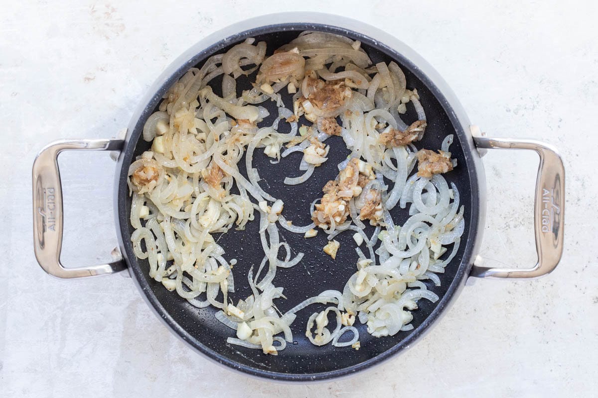 Sliced onions and minced garlic and miso paste sautéing in a black nonstick pan with stainless steel handles on a light surface.