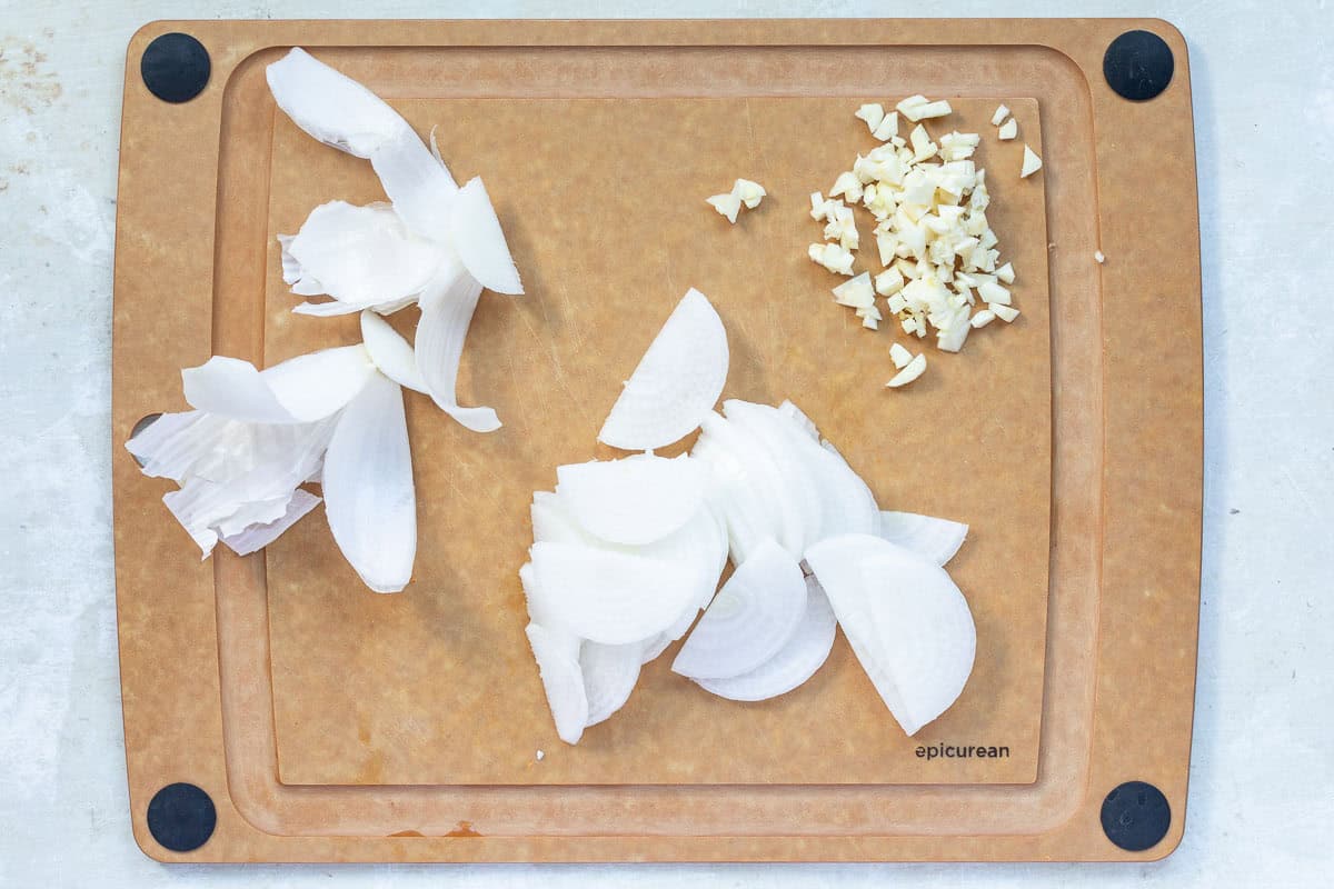 A cutting board with chopped garlic, sliced white onion, and onion peels arranged separately.