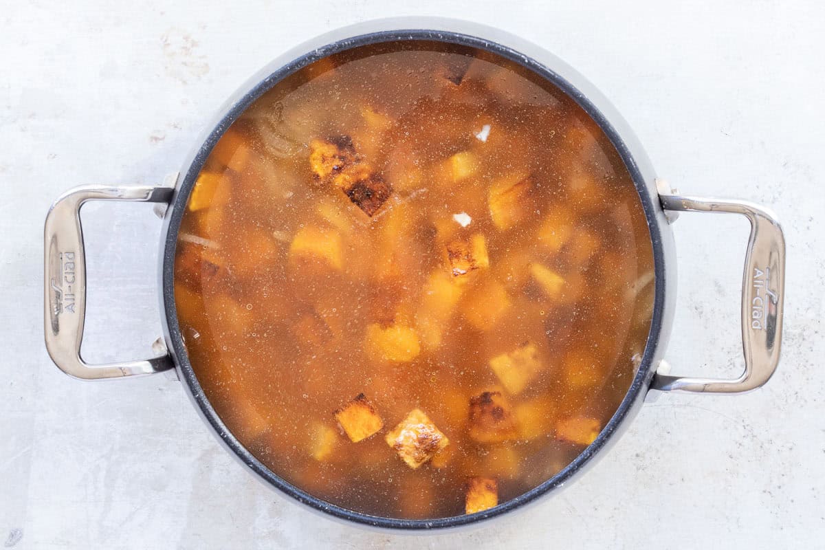 A large pot filled with chunks of roasted butternut squash in broth, viewed from above on a light surface.