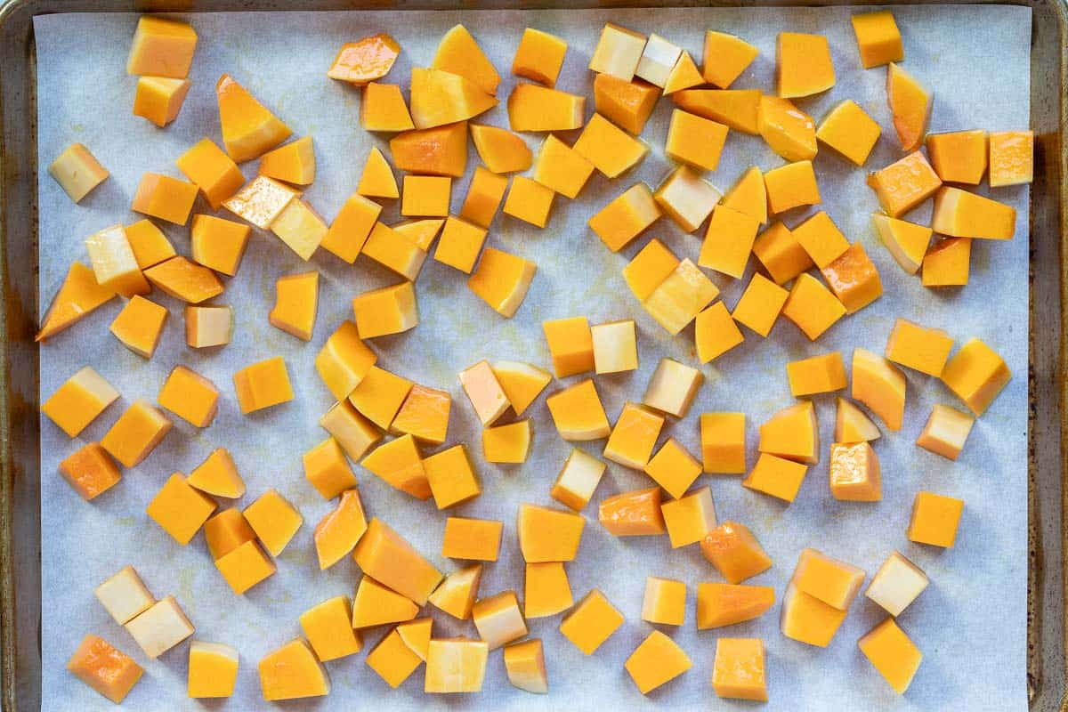 Cubed pieces of raw butternut squash are spread evenly on a parchment-lined baking sheet.
