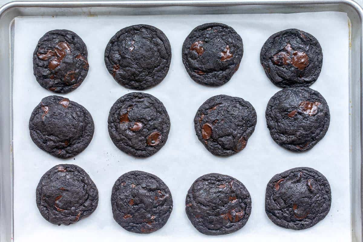 Twelve black cocoa cookies with visible chocolate chunks are arranged in rows on a parchment-lined baking sheet.