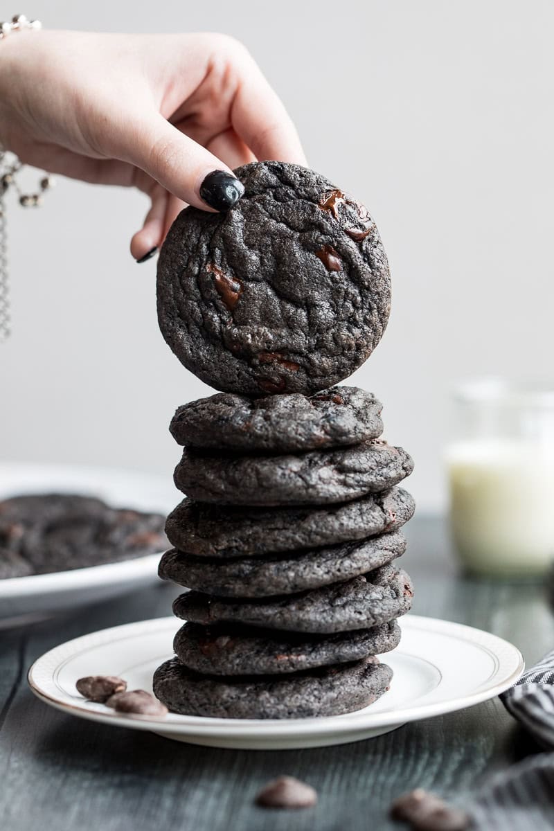 A hand places a black cocoa cookie on top of a stack of similar cookies on a white plate, with milk and more cookies in the background.