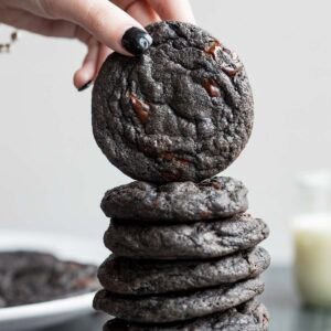 A hand with black nail polish places a black cocoa cookie on top of a stack of similar cookies, with a glass of milk blurred in the background.