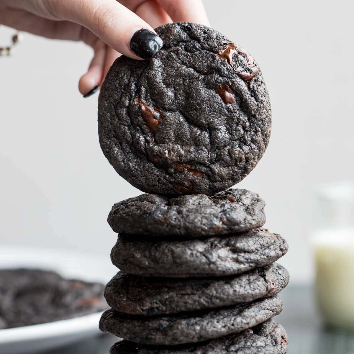 A hand with black nail polish places a black cocoa cookie on top of a stack of similar cookies, with a glass of milk blurred in the background.