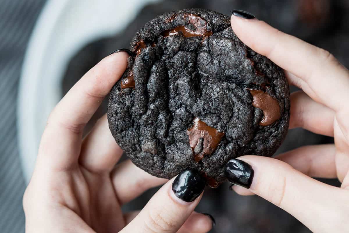 A person with black nail polish holds a dark black cocoa cookie with visible chocolate chunks.