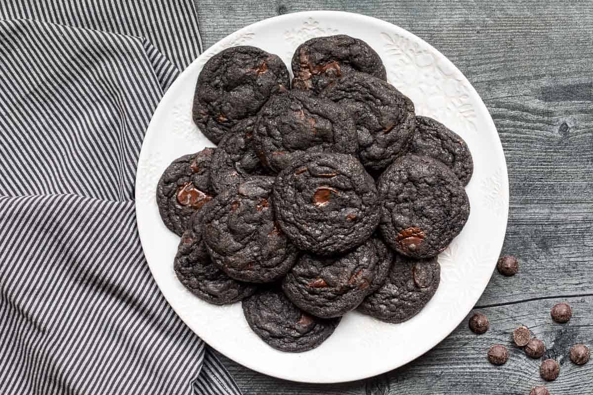 A white plate filled with black cocoa cookies sits on a gray wooden surface near a striped towel and a few scattered chocolate chips.
