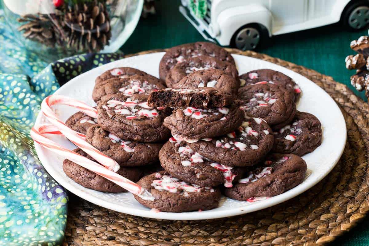 A plate of chocolate peppermint cookies with crushed peppermint topping, garnished with mini candy canes, sits on a woven placemat next to holiday decorations and a white toy truck.