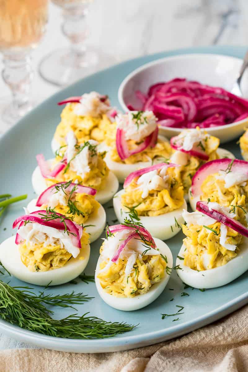 A plate of crab deviled eggs garnished with dill and pickled red onions, with extra pickled onions in a small bowl nearby.