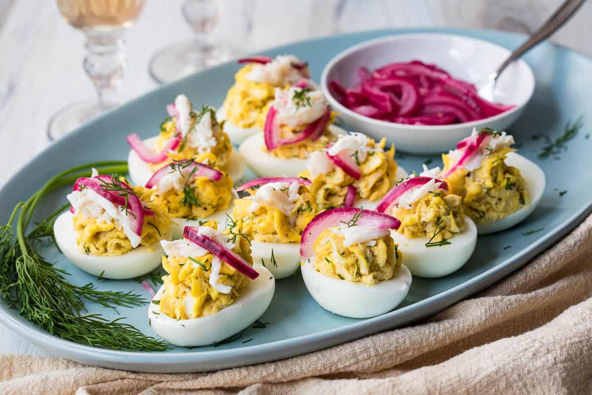 A platter of deviled eggs topped with shredded crab, pickled red onions, and dill, with a bowl of extra pickled onions on the side.