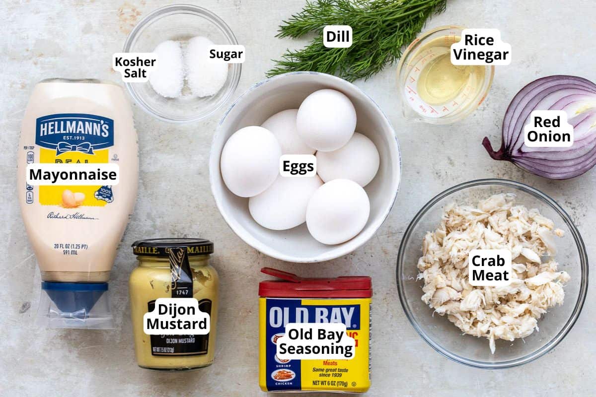 Ingredients for crab deviled eggs and pickled onions, including eggs, mayonnaise, Dijon mustard, Old Bay seasoning, crab meat, red onion, rice vinegar, dill, kosher salt, and sugar, arranged on a countertop.