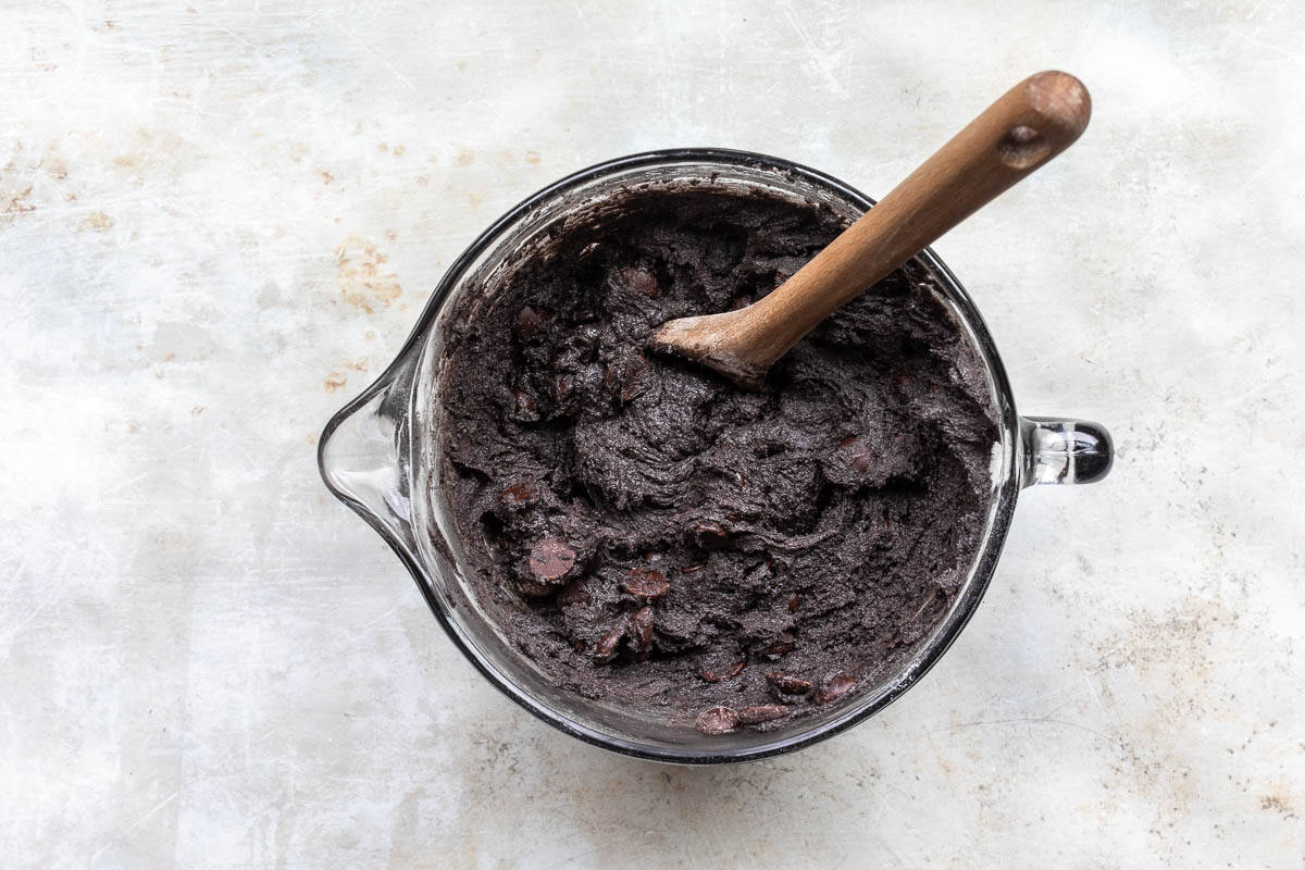 A glass mixing bowl filled with black cocoa cookie batter and a wooden spoon resting inside.