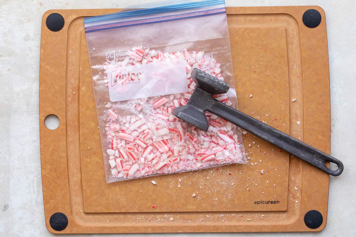 A plastic bag of crushed peppermint candy canes sits on a cutting board with a metal mallet placed on top.