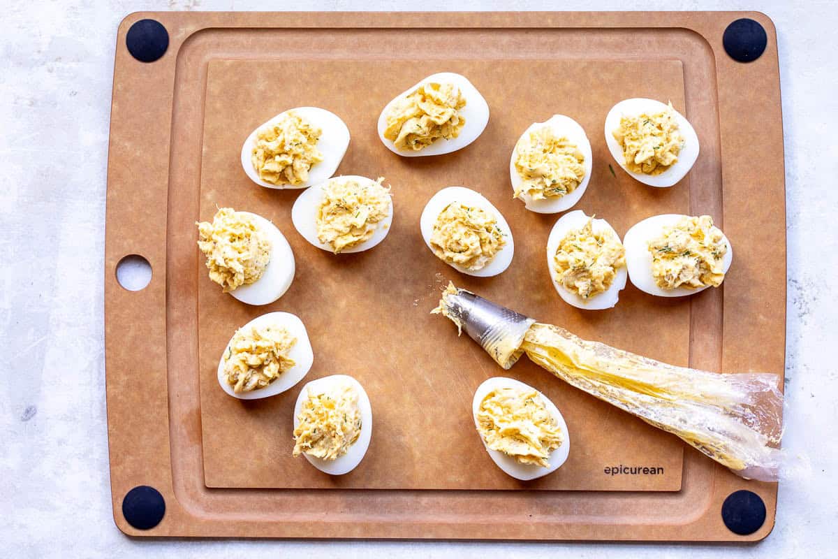 Crab deviled eggs arranged on a brown cutting board, with a piping bag partially filled with yolk mixture beside them.