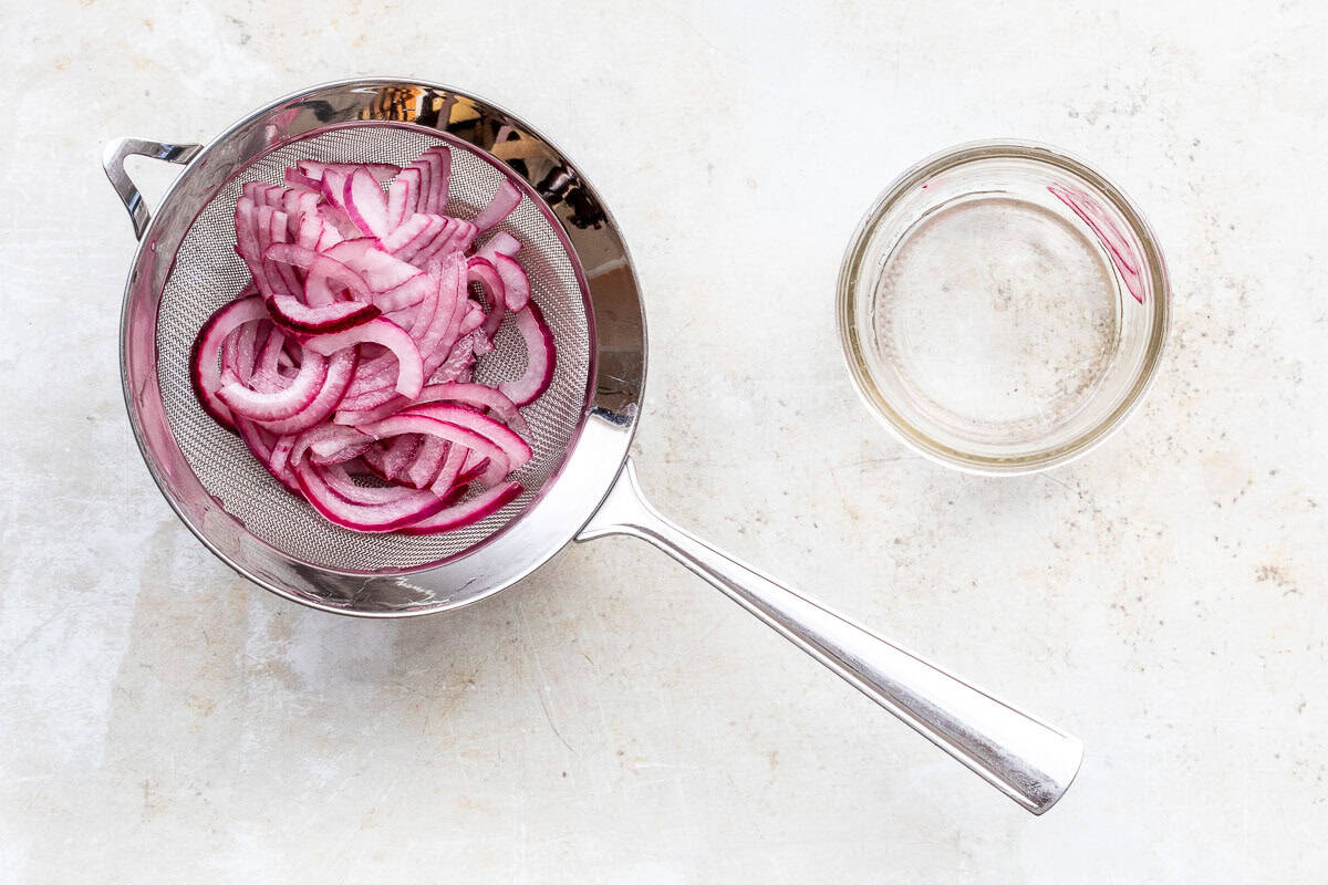 Thinly sliced red onions sit in a metal strainer next to a small mason jar on a light-colored surface.
