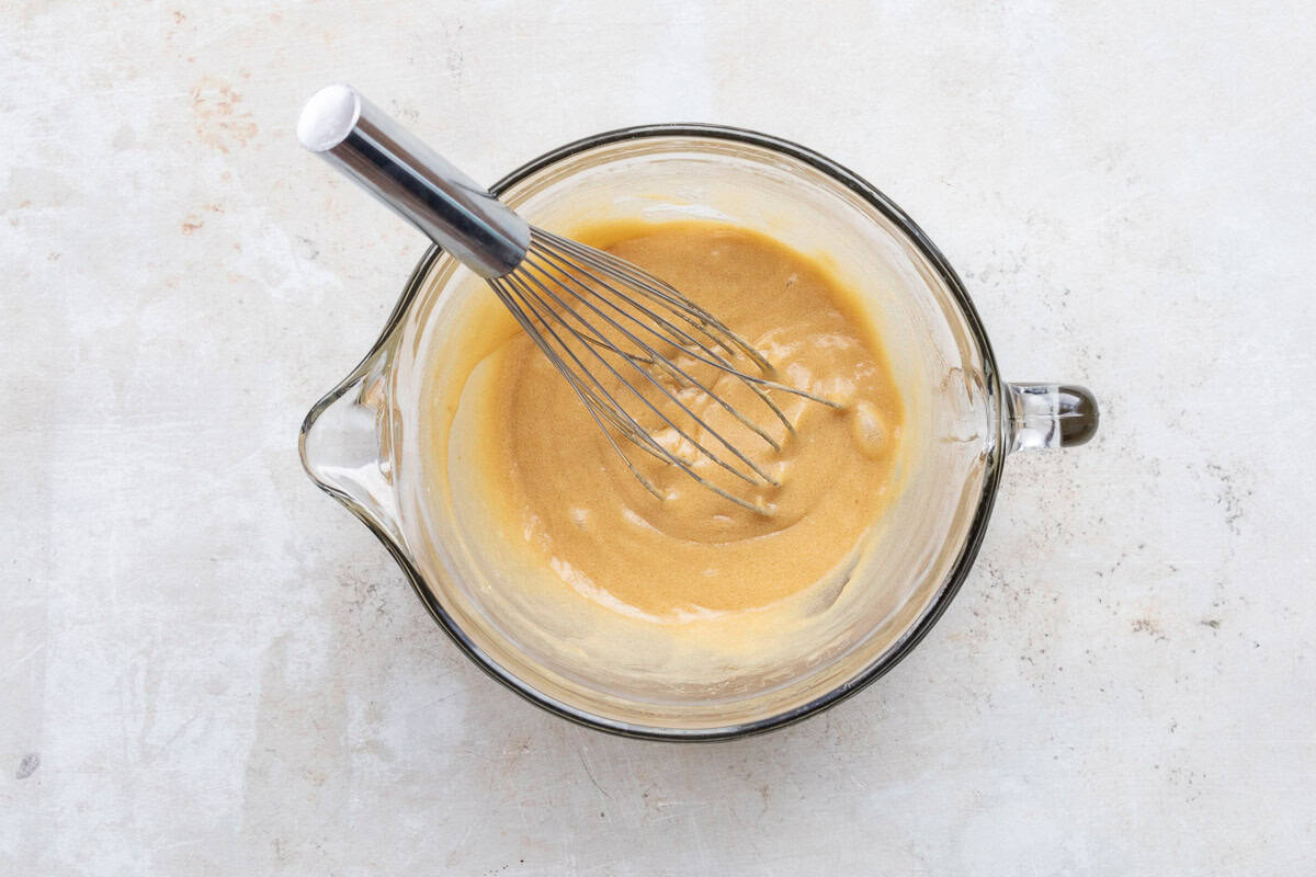 A metal whisk rests in a glass mixing bowl filled with a mixture of eggs, butter, sugar, and brown sugar.