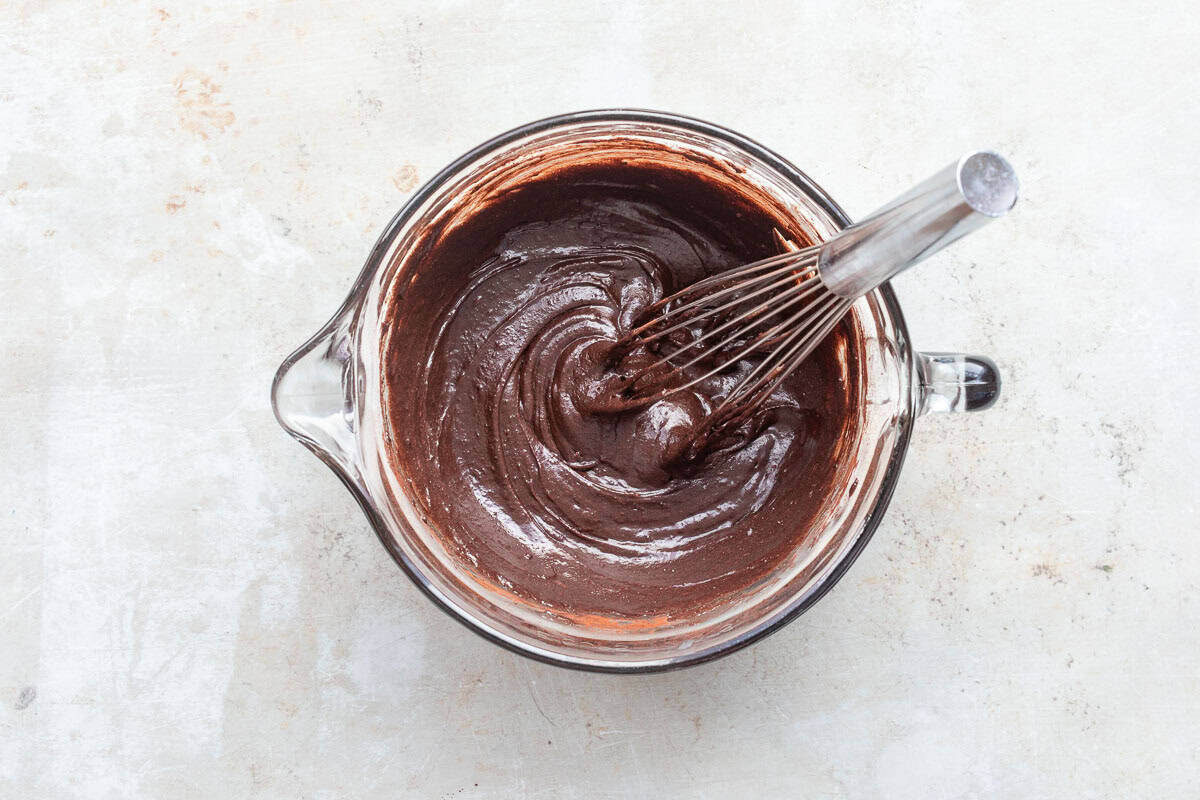 A glass bowl filled with chocolate batter being mixed with a metal whisk, placed on a light surface.