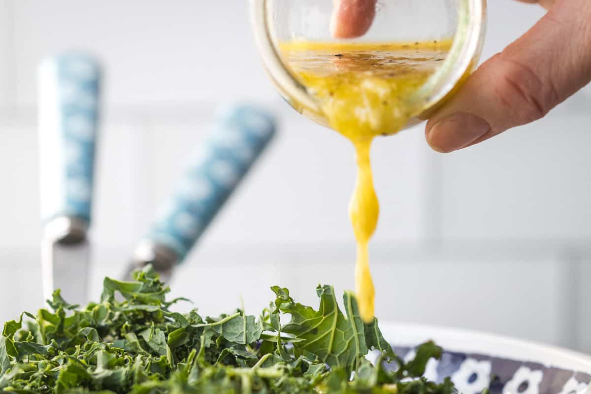 A hand pours kale salad dressing from a small glass container onto fresh green kale in a bowl, with blue-handled utensils in the background.