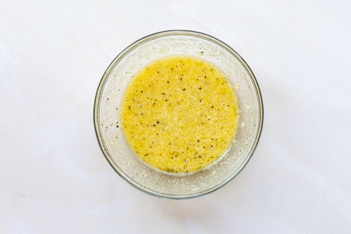 A glass bowl containing a homemade kale salad dressing sits on a light-colored surface.