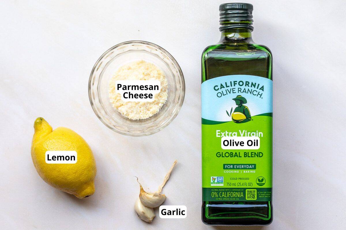 A bottle of extra virgin olive oil, a whole lemon, two garlic cloves, and a bowl of grated Parmesan cheese are arranged on a white surface. Labels identify each item.