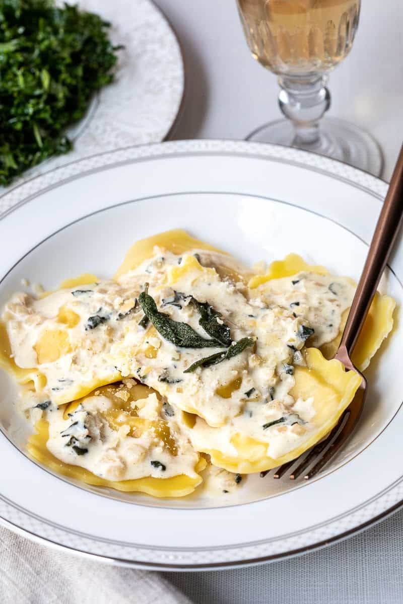 A white plate of lobster ravioli topped with creamy sauce, grated cheese, and sage leaves, with a fork and a glass of white wine nearby.