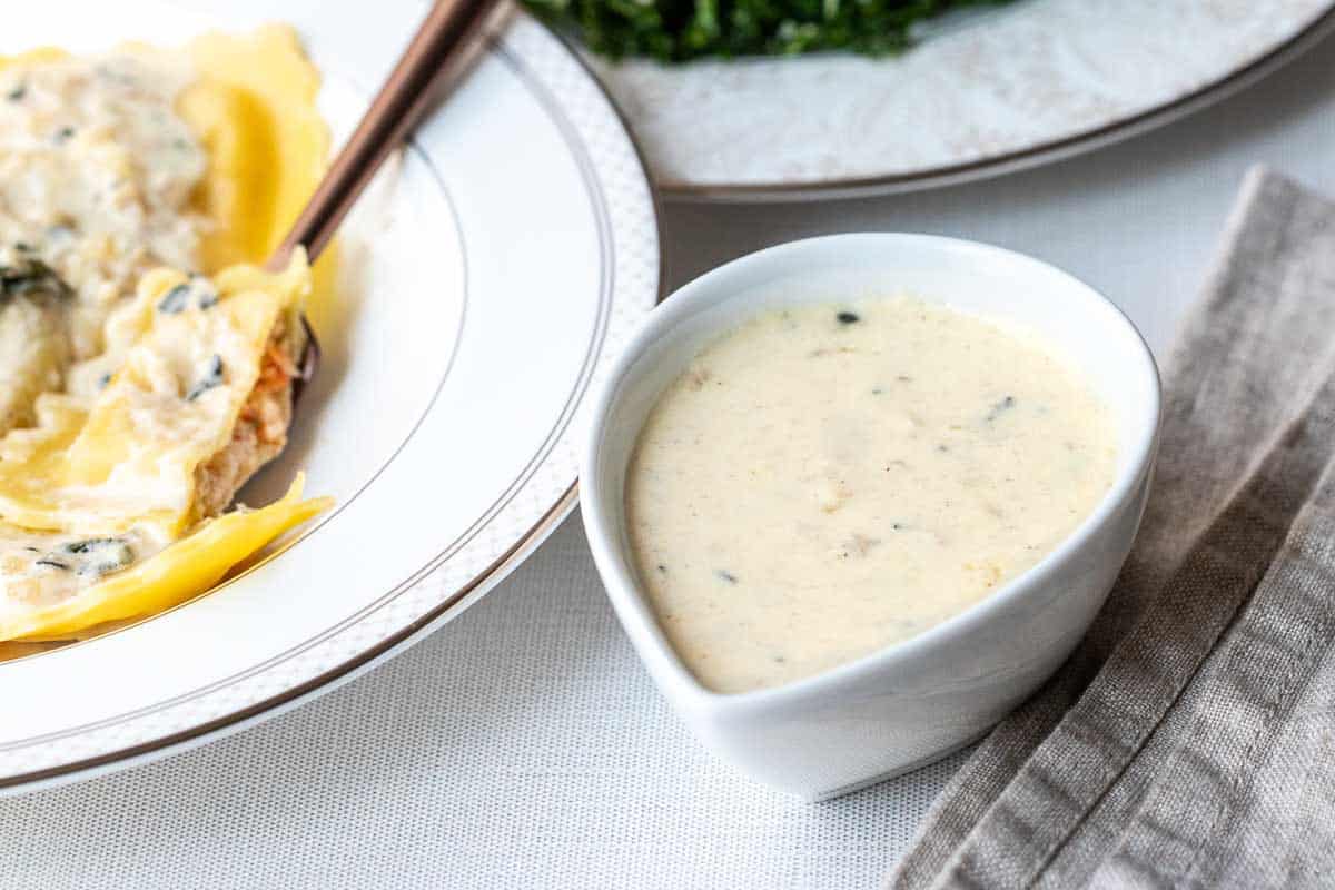 A small white bowl filled with creamy white sauce sits beside a plate of food and a beige striped napkin on a white tablecloth.