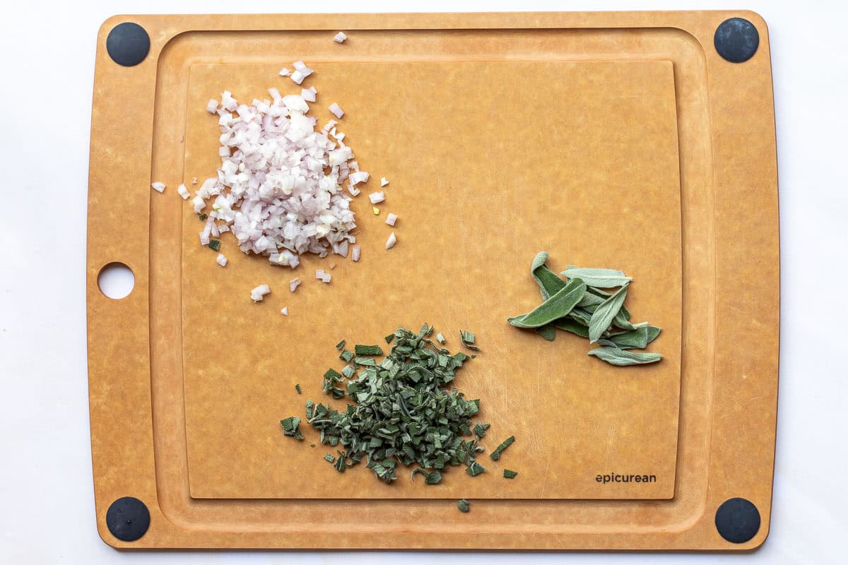 A cutting board with chopped shallots, chopped sage, and whole sage leaves arranged separately on its surface.
