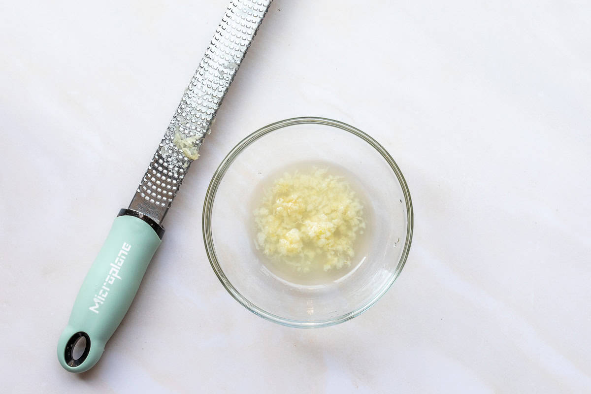 A metal grater with a green handle sits next to a glass bowl filled with freshly grated garlic and lemon juice on a white surface.