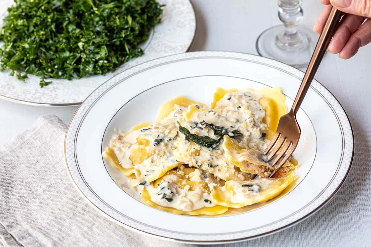 A plate of lobster ravioli with sauce and sage, with a fork taking a bite; a side of chopped greens is visible in the background.