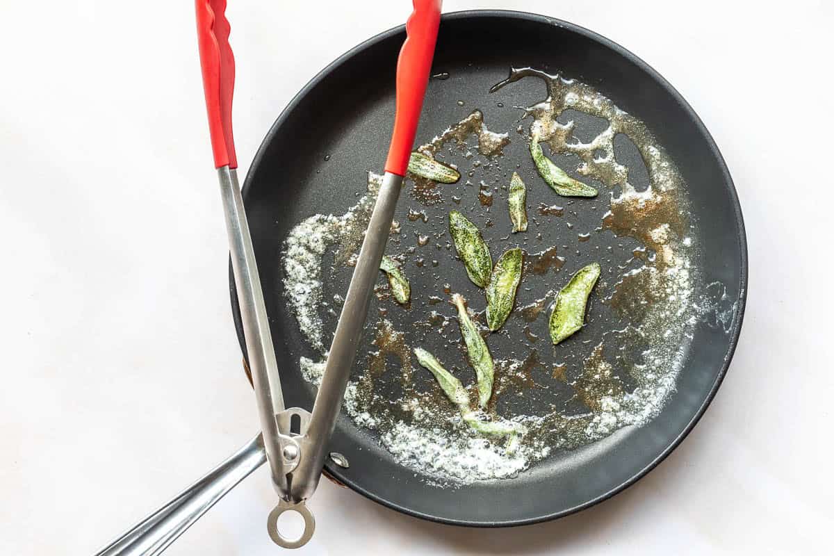 Nonstick frying pan with melted butter, browned bits, and a few fried sage leaves, with red-handled tongs resting on the edge.