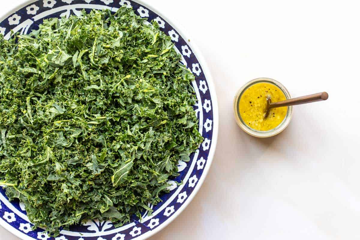 A plate of chopped kale sits beside a small jar of kale salad dressing with a spoon, on a white background.