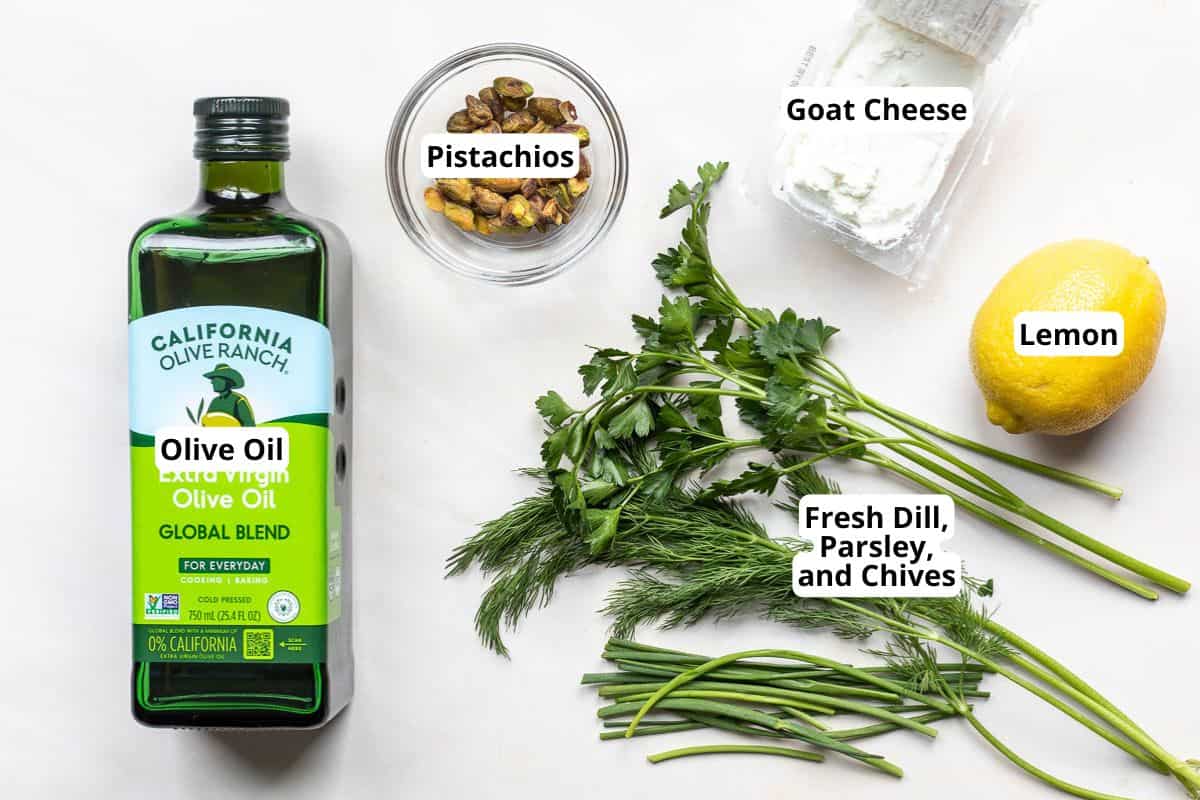 A bottle of olive oil, a bowl of pistachios, a package of goat cheese, a lemon, and fresh dill, parsley, and chives arranged on a white surface.