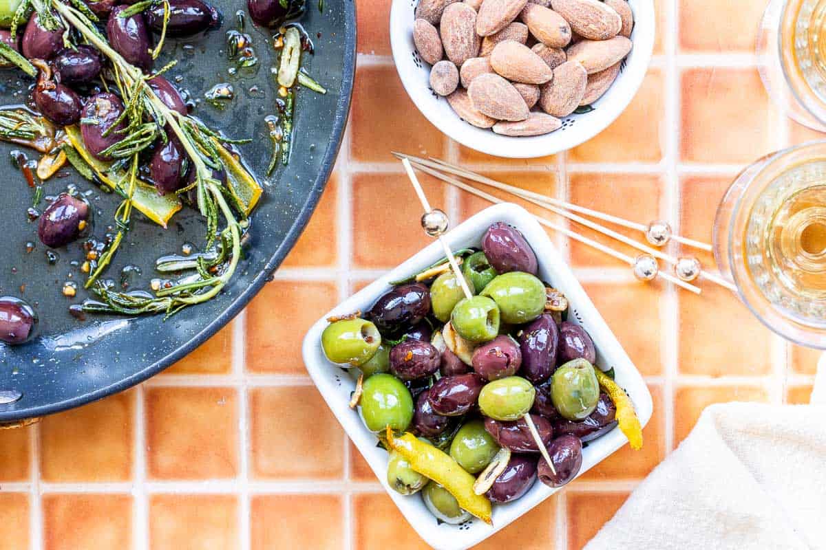 A bowl of mixed marinated olives, a dish of almonds, and a skilled with marinated olives with herbs on an orange tiled surface.