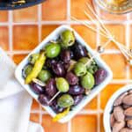 A small white bowl filled with marinated green and black olives, garnished with lemon peel, with toothpicks and almonds nearby on an orange tiled surface.