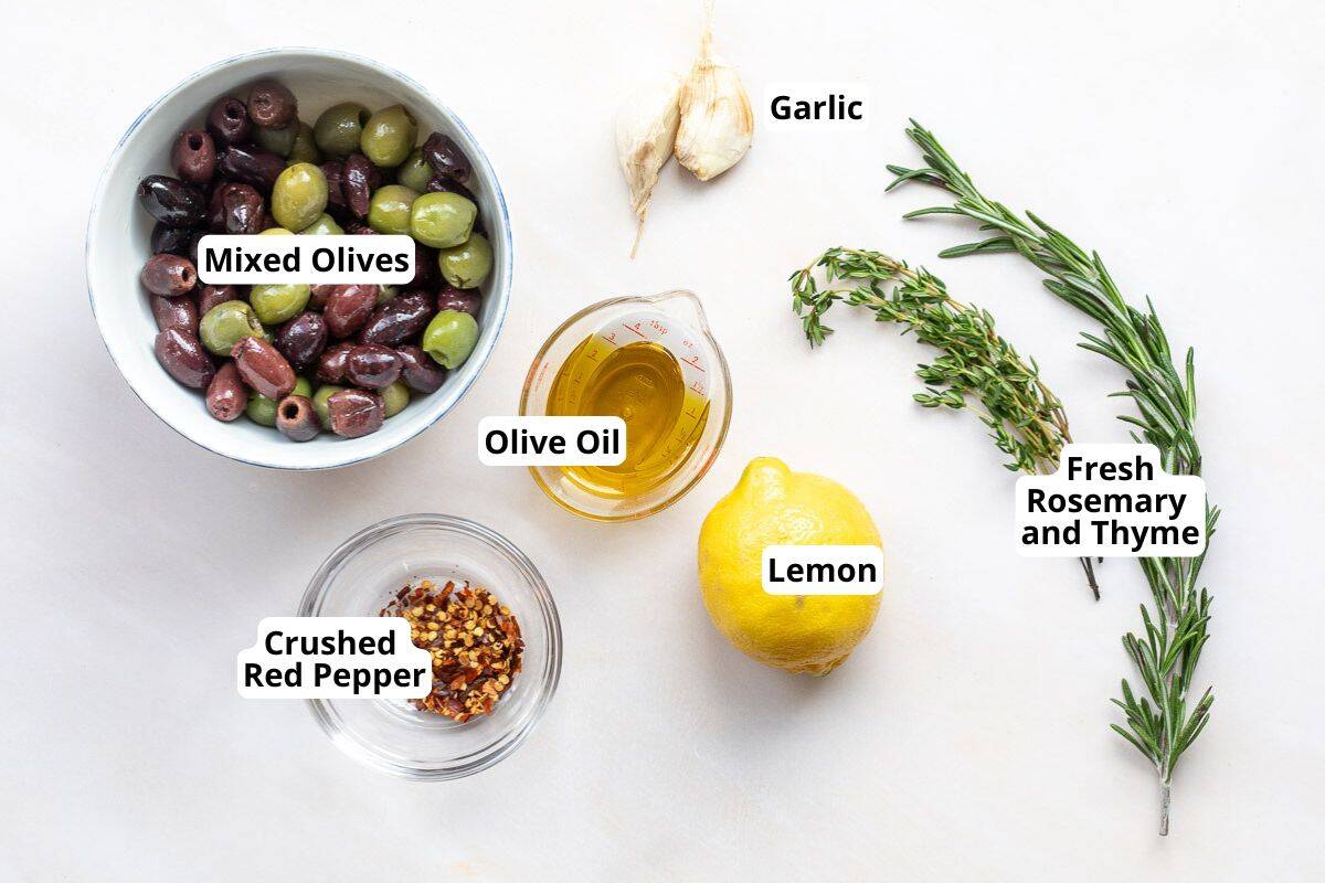 A bowl of mixed olives, olive oil in a glass container, a lemon, garlic cloves, fresh rosemary and thyme, and crushed red pepper displayed on a white surface.
