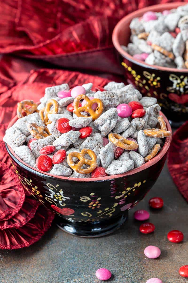 A decorative bowl filled with powdered chocolate snack mix (Valentine's Day muddy buddies), pretzels, and red and pink candy-coated chocolates, with a red fabric background.