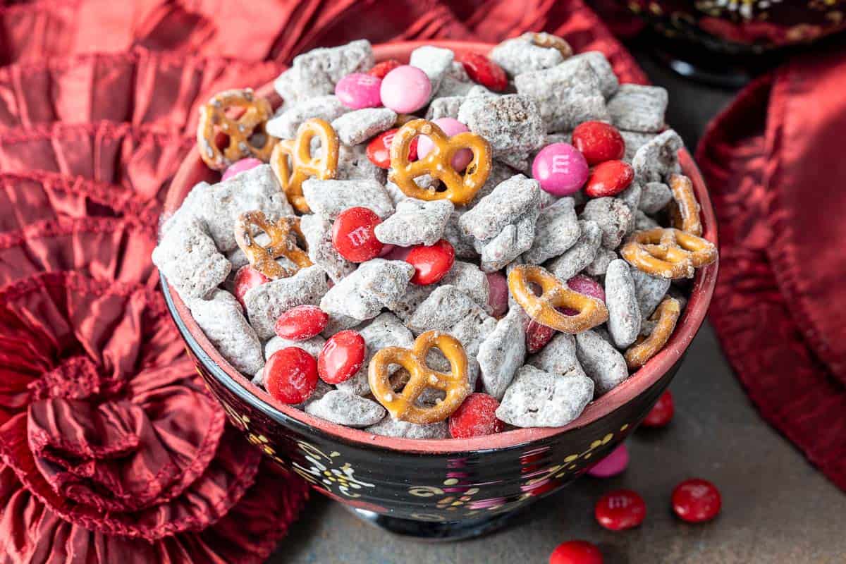 A bowl filled with valentine's puppy chow (chocolate-covered cereal pieces, mini pretzels, and red and pink M&M candies), with red fabric in the background.