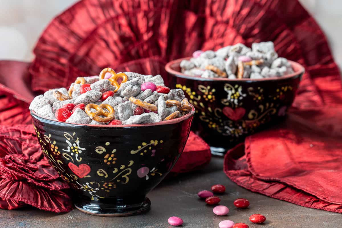 Two black bowls with decorative floral patterns filled with Valentine's Day snack mix including pretzels, coated cereal, and red and pink candies, placed on a red cloth with candies scattered nearby.