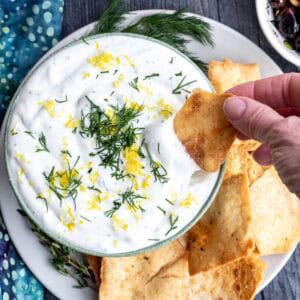 A hand dips a pita chip into a bowl of whipped ricotta dip garnished with dill and lemon zest, with more pita chips on a plate nearby.