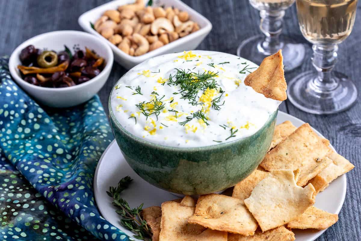 A bowl of creamy whipped ricotta cheese dip topped with dill and lemon zest is served with crackers on a plate, alongside small bowls of nuts and olives, with two glasses of white wine in the background.