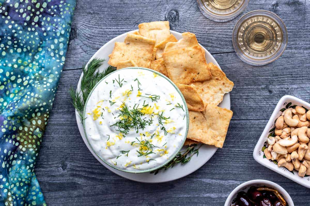 A bowl of creamy whipped ricotta dip topped with herbs and lemon zest, served with crackers on a plate, next to glasses of white wine, nuts, and olives on a wooden table.