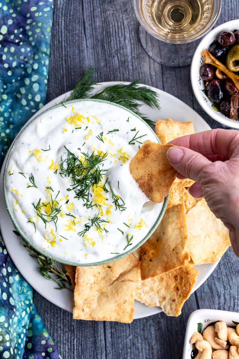 A hand dips a cracker into a bowl of creamy whipped ricotta with goat cheese dip garnished with dill and lemon zest, surrounded by more crackers on a plate. A glass of white wine and small bowls sit nearby.