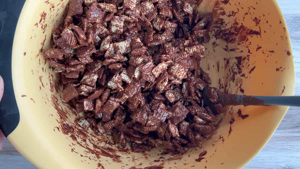 A mixing bowl filled with chocolate-coated cereal pieces being stirred with a spatula.