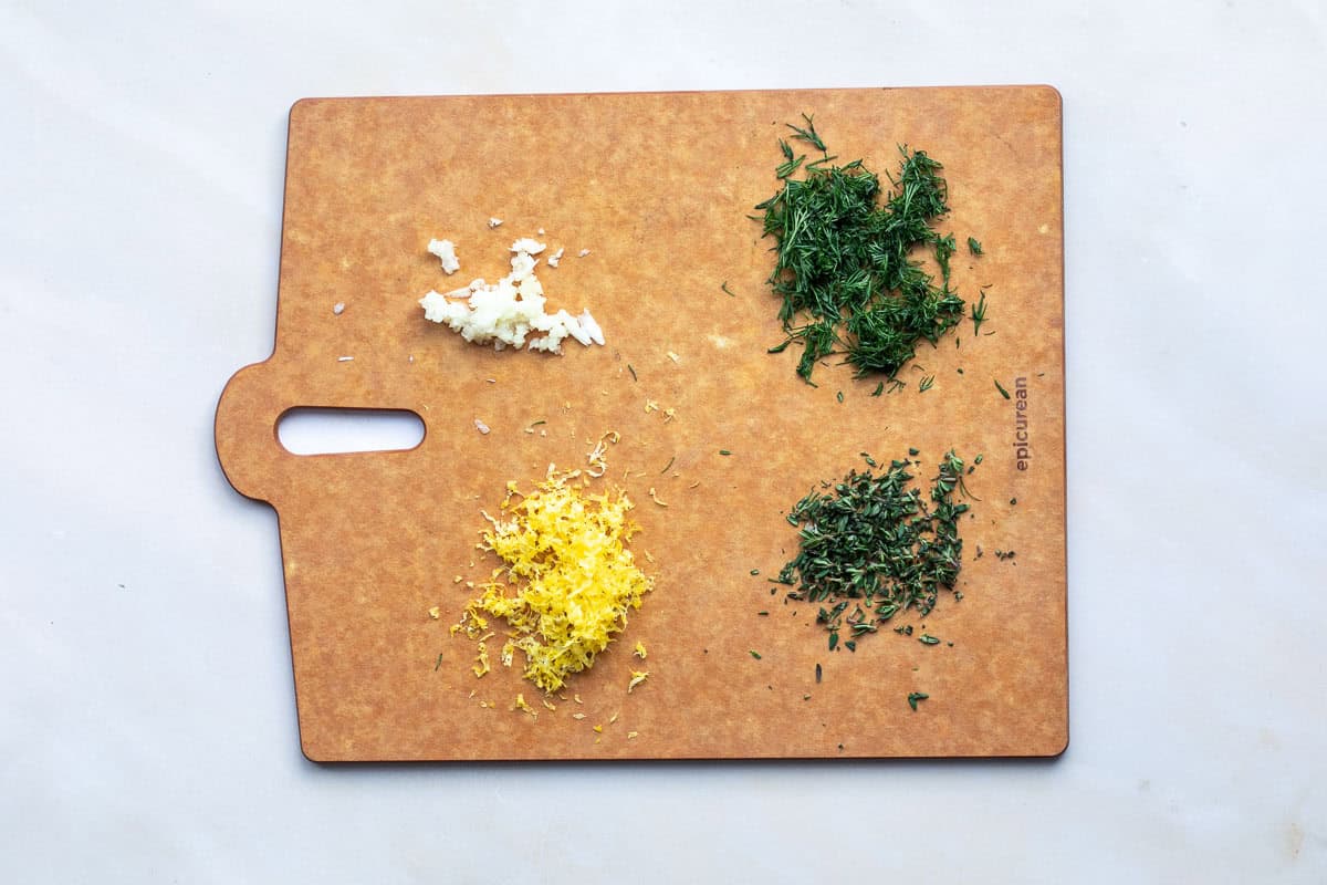 A cutting board with four piles of ingredients: minced garlic, chopped dill, chopped thyme, and lemon zest, arranged separately on a light surface.