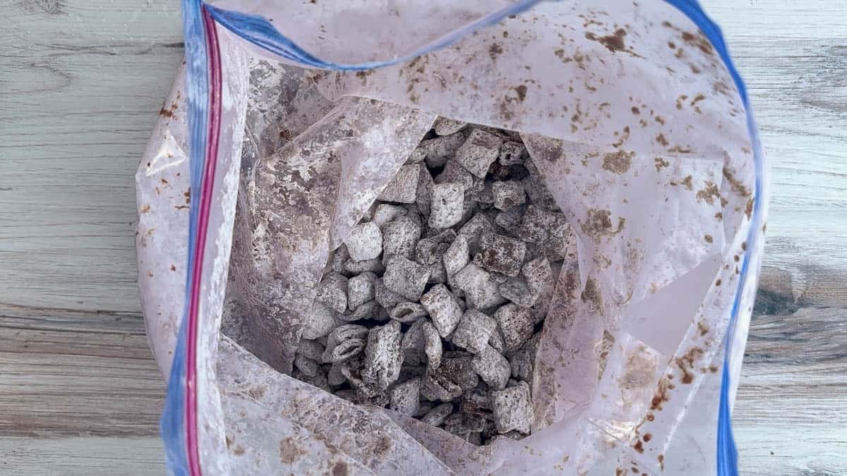 A plastic zip-top bag containing chocolate-coated, powdered sugar-covered cereal snack pieces on a wooden surface.