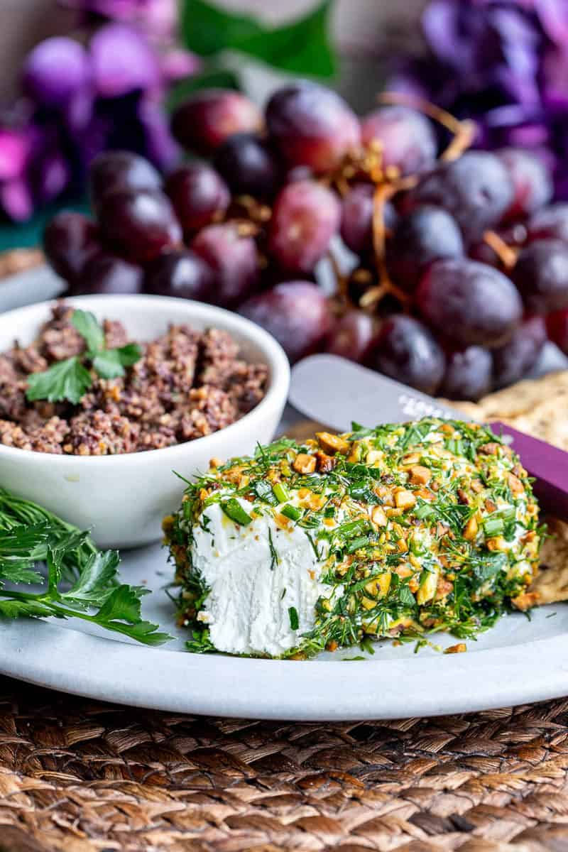 A plate with a herb and nut-coated cheese log, a bowl of tapenade, red grapes, crackers, and fresh herbs.